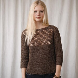 Könnte beinhalten: Ein brauner Pullover mit Dreiviertelärmeln und einem gehäkelten Blumendesign auf der Brust. Der Pullover hat einen runden Ausschnitt und ist aus einem strukturierten Strickstoff gefertigt. Schlichtes Design.