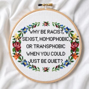 Könnte beinhalten: Ein runder Holzstickrahmen mit weißem Aida-Stoff, der den in schwarzem Kreuzstich gestickten Text enthält: WHY BE RACIST, SEXIST, HOMOPHOBIC, OR TRANSPHOBIC WHEN YOU COULD JUST BE QUIET? Der Text ist von einem Blumenkranz aus roten, blauen und orangefarbenen Blüten umgeben.