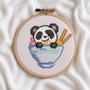 May include: A cross-stitch embroidery piece in a circular wooden hoop featuring a panda bear sitting inside a light blue bowl. The panda has black ears, eye patches, and paws, with pink inner ears. Two yellow chopsticks are placed in the bowl.