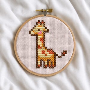 Könnte beinhalten: Ein Kreuzstich-Stickprojekt mit einem verpixelten Giraffenmotiv in Gelb-, Orange- und Brauntönen mit einer rosa Schnauze. Die Stickerei ist in einem runden Holzstickrahmen auf einem hellrosa Stoffhintergrund eingespannt.