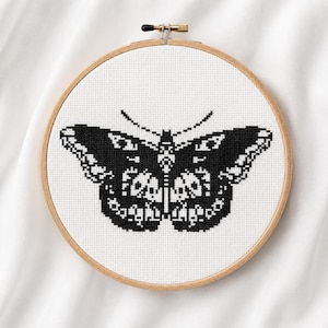 May include: A circular wooden embroidery hoop containing a white fabric panel with a black cross-stitch butterfly design. The butterfly features intricate geometric patterns in black and white, centered within the frame with a metal tension screw.
