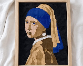 May include: A cross-stitch embroidery of a person wearing a blue headscarf and a large white earring, set against a solid black background. The artwork is encased in a light-colored wooden frame, showcasing a pixelated interpretation of a classic portrait.