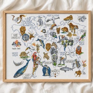 Op de afbeelding: Een ingelijste kruissteek wereldkaart met diverse dieren en geografische labels. Het ontwerp bevat illustraties van een walvis, beer, wolf, leeuw, olifant, panda en pinguïn, met tekst die continenten en oceanen in zwarte draad identificeert.