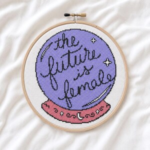Pode incluir: Um bordado em ponto cruz num bastidor de madeira circular com um design de bola de cristal roxa sobre um fundo de tecido branco. O texto "the future is female" está bordado em letras cursivas pretas, acentuado por pequenas estrelas brancas.