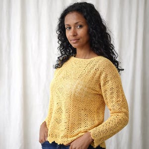 Könnte beinhalten: Ein gelber Häkelpullover mit langen Ärmeln und einem wellenförmigen Saum. Der Pullover hat ein offenes, spitzenartiges Design und einen runden Ausschnitt. Das Model trägt eine blaue Jeans. Der Hintergrund ist ein weißer Vorhang.