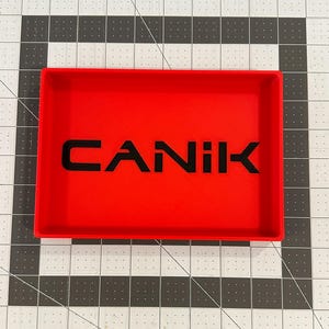 May include: A rectangular red tray with raised edges featuring the word CANIK embossed in black lettering across the center. The tray has a smooth, flat base and is positioned on a white surface with a black grid pattern printed on it.