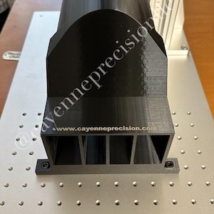 May include: A black 3D printed part with a curved top and a rectangular base with three slots. The part is sitting on a metal surface with a grid pattern. The text "www.cayenneprecision.com" is printed on the base of the part.