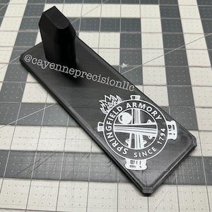 May include: A black rectangular stand with a raised, textured section and the text "@cayenneprecisionllc". The stand features a white logo with the words "SPRINGFIELD ARMORY SINCE 1794".
