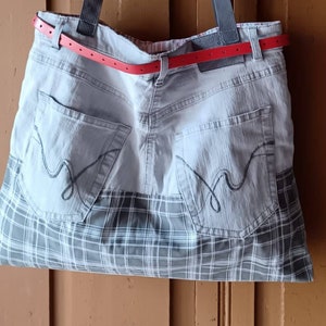 Könnte beinhalten: Eine graue und weiße karierte Tragetasche aus recyceltem Denim mit einem roten Ledergürtel und schwarzen Trägern.