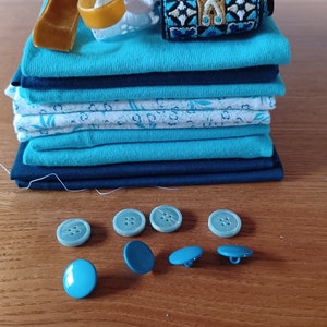 Könnte beinhalten: Ein Stapel Stoff in verschiedenen Blautönen und Weiß, darunter ein gemusterter Stoff, ein einfarbiger blauer Stoff und ein weißer Stoff mit Blumenmuster. Auf einer Holzoberfläche liegen auch ein paar blaue Knöpfe.