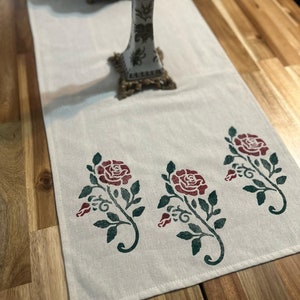 May include: A white table runner with a green and red floral design. The runner features three stylised roses with green leaves and stems. The runner is on a wooden table.