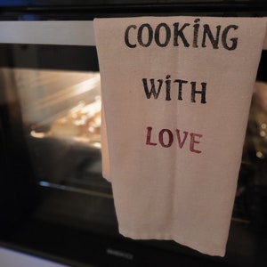 Può includere: Un canovaccio da cucina beige con le parole "COOKING WITH LOVE" stampate in nero e bordeaux. L'asciugamano è appeso alla porta di un forno. L'interno del forno è visibile, con cibo all'interno.