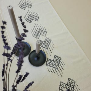 Può includere: Una tovaglia color crema con un motivo geometrico ripetuto in grigio. Due portacandele in metallo nero ciascuno con una candela grigia. Un rametto di lavanda viola è in primo piano.