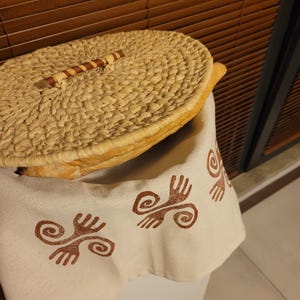 Può includere: Un coperchio di cesto intrecciato con un manico in legno, appoggiato su un tessuto bianco con un motivo a spirale marrone.