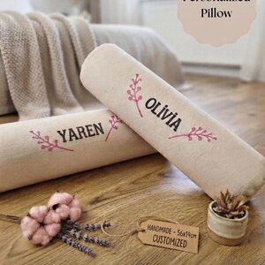 Op de afbeelding: Twee cilindervormige beige kussens met de in het zwart gedrukte namen YAREN en OLIVIA, geaccentueerd door roze takillustraties. Een bruin papieren label toont de tekst HANDMADE - 56x14cm CUSTOMIZED, naast roze katoentakken.