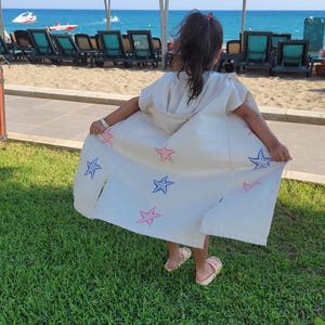 May include: A white hooded beach cover-up with a blue and pink starfish pattern. The cover-up is worn by a person standing on green grass.