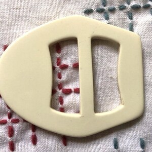 Op de afbeelding: Een witte plastic gesp met twee gleuven voor een riem of band.