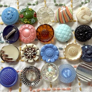 May include: A collection of twenty assorted round buttons arranged in a grid. The buttons feature various colors including blue, green, clear, pink, brown, black, and cream, with diverse textures, patterns, and finishes such as floral, striped, and geometric designs.
