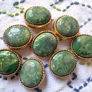 May include: Eight vintage green and gold buttons with a marbled effect. The oval-shaped buttons have a gold rope-like border. The green color varies in shade, creating a unique look. These buttons are perfect for sewing projects or adding a vintage touch to clothing.