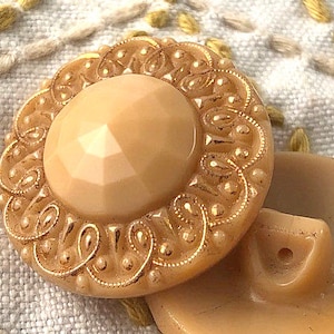 Può includere: Un grande bottone ornato, color crema, con un centro rialzato sfaccettato e un design intricato color oro attorno al bordo.