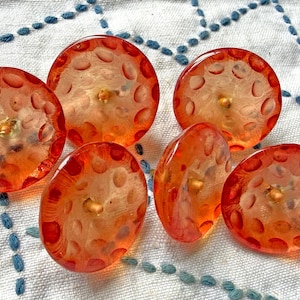 Könnte beinhalten: Sechs orange-rote, runde Knöpfe mit strukturierter Oberfläche und goldfarbenem Zentrum. Die Knöpfe sind auf einem weißen Stoff mit blauen Nähten angeordnet. Diese Knöpfe sind ideal für Näh- und Bastelprojekte.
