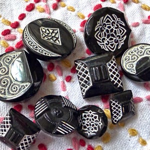 Può includere: Una collezione di nove bottoni neri con vari motivi incisi in bianco, tra cui griglie geometriche, complessi motivi floreali e forme a mezzaluna. I bottoni variano nella forma, con la presenza di design sia circolari che quadrati.