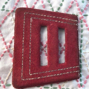 Puede incluir: Un posavasos cuadrado de ante rojo con dos recortes rectangulares y costuras blancas. El posavasos tiene una superficie texturizada. El fondo es un paño blanco con patrones de diamantes bordados en rosa y verde.