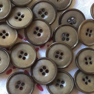 Può includere: Una collezione di bottoni in plastica marrone con quattro fori ciascuno. I bottoni sono disposti in modo casuale su una superficie di tessuto bianco.