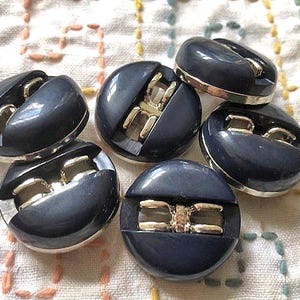Puede incluir: Un conjunto de seis botones azul marino con un acento metálico plateado. Los botones tienen un diseño único con una forma redondeada y una ranura horizontal. Los botones están dispuestos sobre una tela blanca con costuras coloridas.