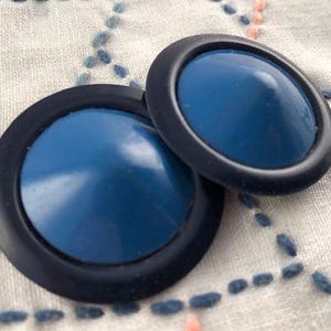 Op de afbeelding: Twee ronde knopen met een glad, gewelfd blauw midden omringd door een platte, matzwarte buitenrand. De knopen hebben een verhoogd, conisch profiel en een eenvoudig ontwerp, wat een duidelijk contrast laat zien tussen de blauwe en zwarte kleuren.