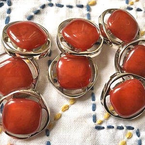 May include: Eight vintage buttons with a reddish-orange, oval stone set in a silver-toned metal frame. The buttons are displayed on white fabric with blue and yellow stitching. The buttons are a classic design.