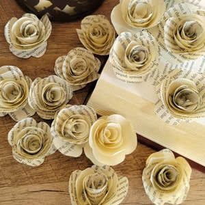 May include: A collection of 10 paper roses made from old book pages. The roses are arranged on a wooden surface with a book and a decorative lamp in the background.