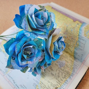 May include: A bouquet of three blue and white paper roses made from maps. The roses are arranged in a cluster with the stems facing down.