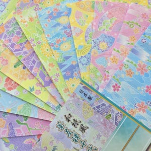 May include: A collection of rectangular sheets of decorative paper featuring various floral and fan patterns in pastel shades of pink, blue, yellow, green, and purple. The paper has a textured surface and includes Japanese text and the phrase 日本製.
