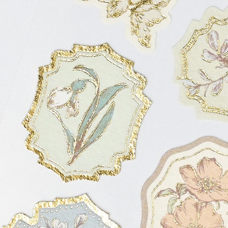 Op de afbeelding: Close-up van verschillende bloemenstickers met gouden folieranden. De stickers bevatten delicate illustraties van bloemen in zachte pastelkleuren, waaronder lichtgroen, blauw en perzik. De ontwerpen staan tegen een cr&egrave;mekleurige achtergrond.