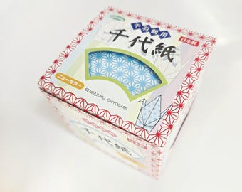 May include: A box of Japanese origami paper, featuring a fan-shaped design with blue and white star patterns. The box has red and white geometric patterns and Japanese text. The text "SENBAZURU CHIYOGAMI" is also visible.