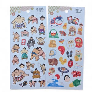 May include: Two sticker sheets featuring Japanese-themed illustrations. One sheet has sumo wrestlers in various poses. The other sheet has a lucky cat, Mount Fuji, and other traditional Japanese symbols. The stickers are on a light blue background.