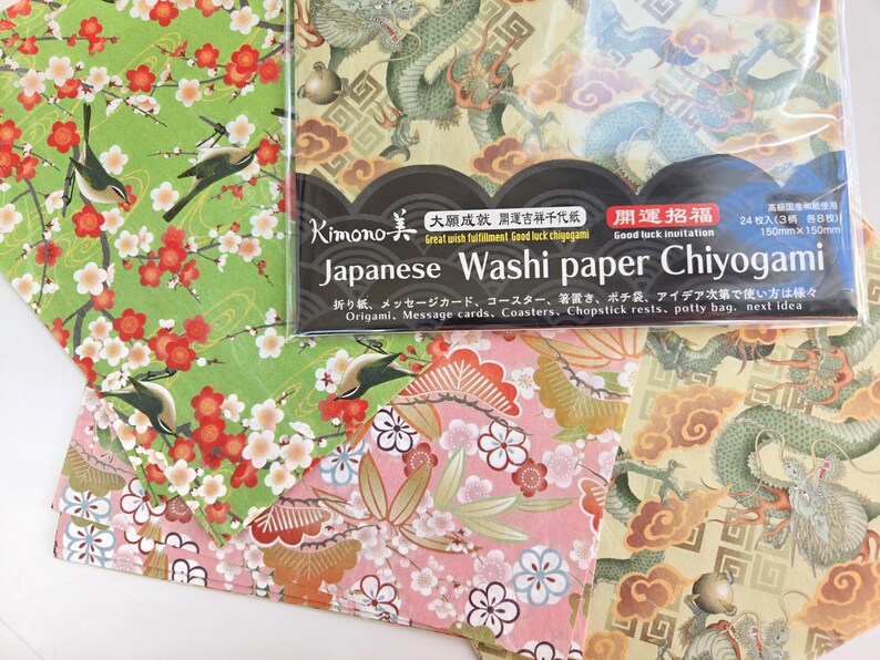May include: A pack of Japanese Washi paper Chiyogami with four different designs. The designs include a green background with white flowers and birds, a pink background with white flowers and gold accents, a yellow background with a green dragon, and a yellow background with a green dragon and gold accents. The pack includes 24 sheets of paper, each measuring 150mm x 150mm. The text on the pack reads "Kimono 大成運吉祥千代紙 Great wish fulfillment Good luck chayogami 開運招福 Good luck invitation Japanese Washi paper Chiyogami 折り紙、メッセージカード、コースター、箸置き、ポチ袋、アイデア次第で使い方は様々 Origami, Message cards, Coasters, Chopstick rests, potty bag, next idea"