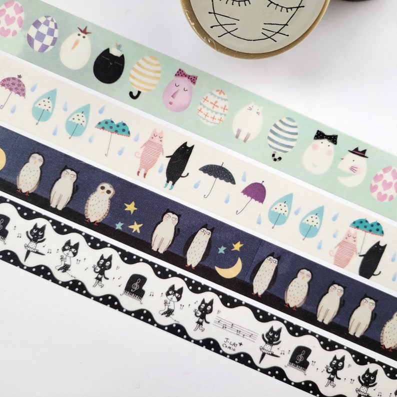 May include: Four rolls of washi tape with different designs. The first roll has a green background with white eggs, black cats, and pink faces. The second roll has a white background with blue raindrops, umbrellas, and pink cats. The third roll has a dark blue background with white owls, yellow stars, and a crescent moon. The fourth roll has a black and white background with black cats playing musical instruments.