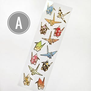 K&ouml;nnte beinhalten: Ein Bogen mit Origami-Kranich-Aufklebern in verschiedenen Farben und Mustern, darunter Blumen- und geometrische Designs. Die Kraniche sind in Blau-, Orange-, Gr&uuml;n-, Rosa- und Goldt&ouml;nen gehalten und haben einen metallischen Glanz. Ein grauer Kreis mit dem Buchstaben "A" befindet sich in der oberen linken Ecke.