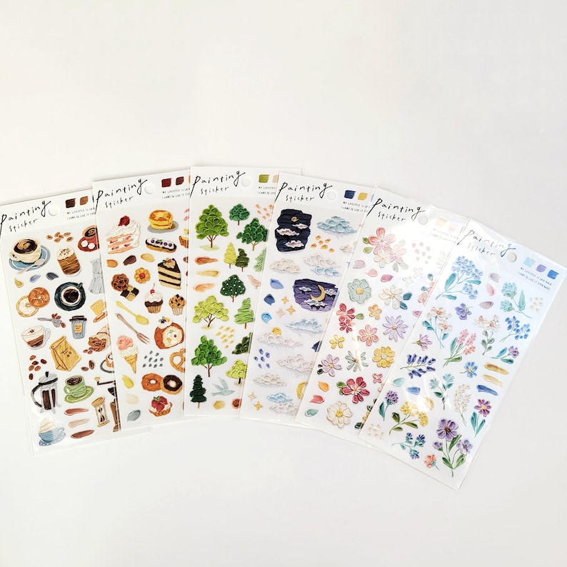 Op de afbeelding: Een verzameling sticker vellen met diverse ontwerpen. De vellen bevatten afbeeldingen van koffie, gebak, bomen, wolken, bloemen en een nachtsc&egrave;ne met een maan. Op elk vel staat het woord "Painting Sticker".