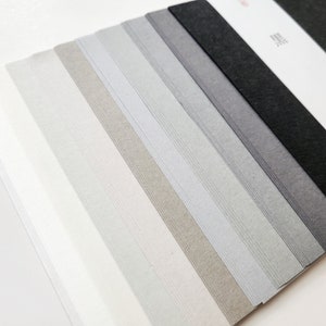 May include: A collection of paper sheets in varying shades of gray, from light to dark, arranged in a diagonal pattern. Each sheet has subtle, parallel lines. The top right corner shows a white sheet with Japanese characters.