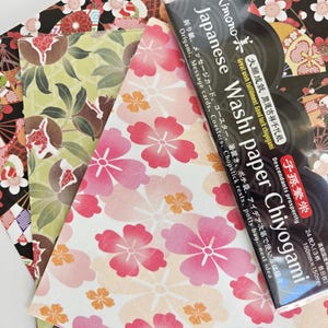 May include: A pack of Japanese Washi paper Chiyogami with three different designs. The designs include a black and white floral pattern, a green and brown floral pattern, and a pink and white floral pattern. The pack includes 24 sheets of paper, 8 sheets of each design. The paper is 150mm x 150mm.