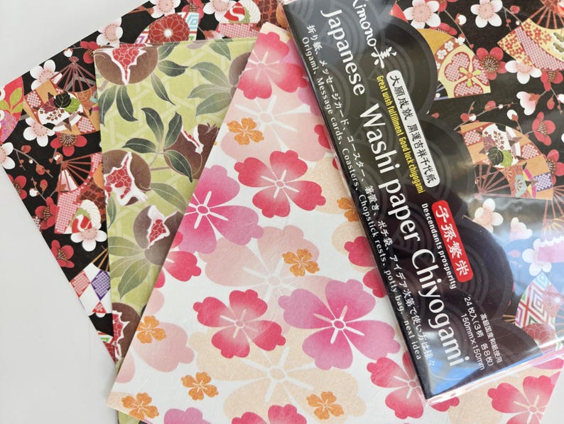 May include: A pack of Japanese Washi paper Chiyogami with three different designs. The designs include a black and white floral pattern, a green and brown floral pattern, and a pink and white floral pattern. The pack includes 24 sheets of paper, 8 sheets of each design. The paper is 150mm x 150mm.