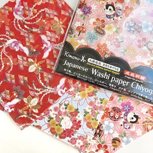 May include: A pack of Japanese Washi paper Chiyogami, featuring three different designs. The designs include a red background with white cranes, a blue background with pink and yellow flowers, and a pink background with a white cat and flowers. The pack includes 24 sheets of paper, each measuring 15cm x 15cm. The text on the pack reads "Kimono Japanese Washi paper Chiyogami" and "Great with fulfillment, Good luck charms".