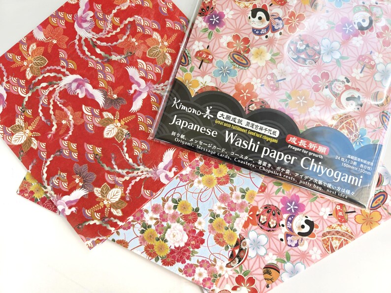 May include: A pack of Japanese Washi paper Chiyogami, featuring three different designs. The designs include a red background with white cranes, a blue background with pink and yellow flowers, and a pink background with a white cat and flowers. The pack includes 24 sheets of paper, each measuring 15cm x 15cm. The text on the pack reads "Kimono Japanese Washi paper Chiyogami" and "Great with fulfillment, Good luck charms".