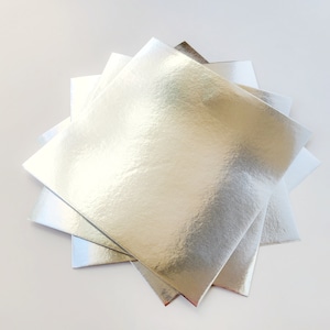 May include: A stack of four silver square cake boards. The boards are made of a shiny, metallic material.