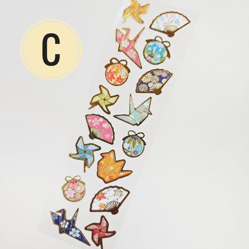 K&ouml;nnte beinhalten: Ein Streifen dekorativer Aufkleber mit verschiedenen japanischen Motiven. Die Aufkleber umfassen Origami-Kraniche, F&auml;cher, Windr&auml;der und runde Ornamente, alle mit Goldfolienakzenten. Der Buchstabe "C" befindet sich in einem gelben Kreis oben links.