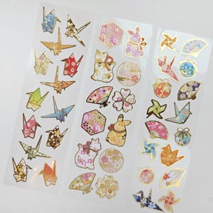 K&ouml;nnte beinhalten: Bogen mit dekorativen Aufklebern mit Origami-Kranichen, F&auml;chern, Hasen und floralen Designs. Die Aufkleber haben goldfarbene Akzente und sind in verschiedenen Farben wie Rosa, Blau und Gold gehalten. Ideal f&uuml;r Scrapbooking und Bastelarbeiten.