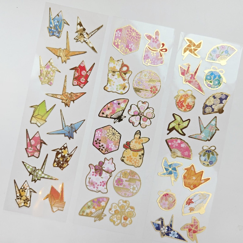K&ouml;nnte beinhalten: Bogen mit dekorativen Aufklebern mit Origami-Kranichen, F&auml;chern, Hasen und floralen Designs. Die Aufkleber haben goldfarbene Akzente und sind in verschiedenen Farben wie Rosa, Blau und Gold gehalten. Ideal f&uuml;r Scrapbooking und Bastelarbeiten.